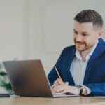 Corporate worker in formal suit sits at desktop focused at laptop computer poses in office interior writes down information with pencil watches tutorial webinar browses website makes research
